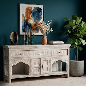 May include: A white-washed wooden sideboard with intricate carvings, arched features, and drawers. The sideboard is decorated with a framed abstract artwork, a vase of dried flowers, and a potted plant, creating a stylish display.