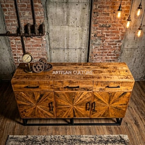 Puede incluir: Aparador rústico de madera con tres puertas y dos cajones, con un diseño de rayos de sol. El mueble está sostenido por un marco de metal negro y tiene tiradores negros. El texto "ARTISAN CULTURE" es visible. El fondo incluye ladrillo y hormigón.