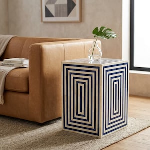 Può includere: Un tavolino quadrato con un motivo geometrico blu navy e crema. Il motivo è composto da rettangoli ripetuti. Sopra c'è un vaso di vetro con una foglia verde. Il tavolo è accanto a un divano in pelle marrone. Dimensioni: circa 40 cm x 40 cm.