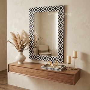 Op de afbeelding: Een rechthoekige spiegel met een zwart-witte geometrische rand hangt boven een houten console. Een vaas met gedroogde planten, een decoratief dienblad en twee kandelaars met kaarsen staan op de tafel.