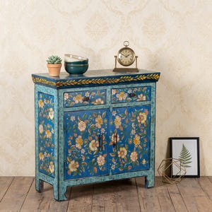 May include: A blue cabinet with floral designs, two drawers, and two doors. The cabinet has a gold trim and is adorned with yellow and white flowers. A small clock, bowls, and a plant sit on top. A framed fern print is on the right.