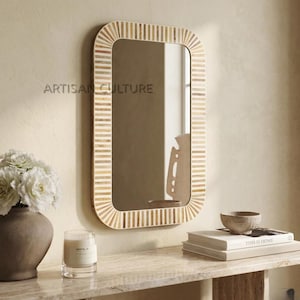 May include: Rectangular mirror with a striped, light-colored frame. The mirror is hanging on a beige wall. The text "ARTISAN CULTURE" is printed on the wall. A vase with white flowers, a candle, and books are on a table below.