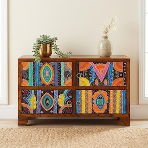 May include: A wooden chest of drawers with four drawers, each decorated with a bright, colourful geometric pattern. The drawers display orange, blue, yellow, and pink designs on a dark background. A small succulent in a gold pot and a vase with dried flowers sit on top.