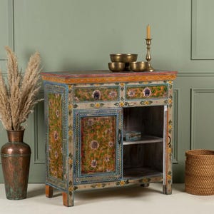 May include: A hand-painted wooden cabinet with floral designs in shades of green, yellow, and blue. The cabinet has two drawers, a single door, and an open shelf. Brass bowls and a candle holder sit on top. A copper vase and a woven basket are on either side.
