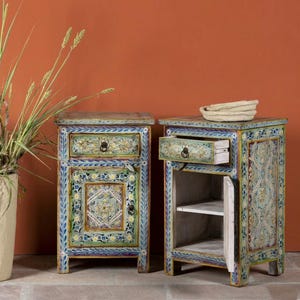 May include: Two hand-painted wooden bedside tables. The tables are painted in shades of blue, green, and yellow with floral and geometric patterns. One drawer is open, revealing the interior. A woven basket sits on top of one table.