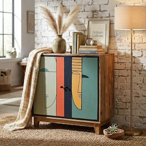 May include: A wooden cabinet with a colorful abstract face design on the doors. The cabinet has a light brown finish and is decorated with a vase of dried plants, books, and a framed print that reads "Artisan Culture".