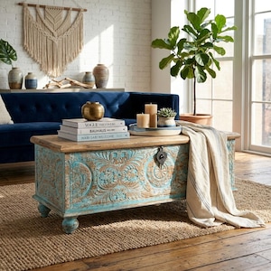 Op de afbeelding: Een distressed turquoise en houten salontafel met gesneden bloemmotieven. De tafel heeft een houten blad en is versierd met boeken, kaarsen en een decoratief dienblad. Een beige plaid met blauwe strepen ligt op de tafel.