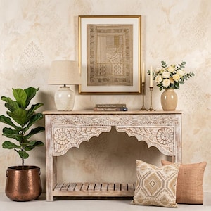 May include: A decorative console table with carved floral details, a framed textile art piece, and a table lamp. The table is adorned with a vase of flowers, candles, and decorative pillows. A potted plant in a copper pot is on the left.