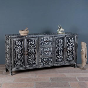 May include: Black and white credenza with ornate floral designs. The cabinet has four doors and three drawers, with a decorative sculpture and mortar and pestle on top. The credenza is made of wood and has a rectangular shape.