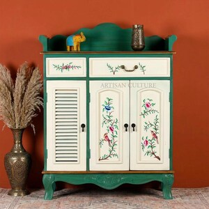 May include: An off-white cabinet with green trim, hand-painted with floral and bird motifs. The cabinet features two doors, a drawer, and a small shelf. The text "ARTISAN CULTURE" is displayed on the doors. A small elephant figurine and a decorative vase are on the shelf.