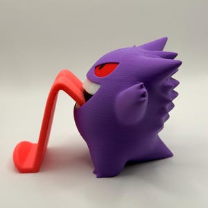 May include: A purple Gengar figurine with red eyes and a white mouth, holding a red phone stand. The figurine is a 3D-printed item, showcasing a popular character from a video game.