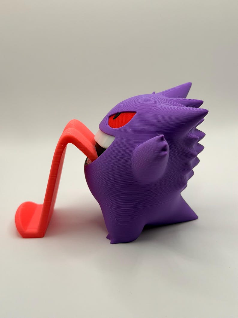 May include: A purple Gengar figurine with red eyes and a white mouth, holding a red phone stand. The figurine is a 3D-printed item, showcasing a popular character from a video game.