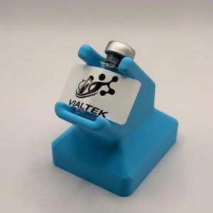 May include: A light blue vial holder with a clear glass vial and a silver cap. The holder has a rectangular base and a hand-shaped top that cradles the vial. A white label with the text "VIALTEK" is attached to the holder.