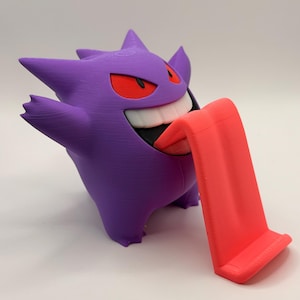 May include: A purple Gengar figure with red eyes and a wide grin, holding out a bright red tongue that serves as a phone or tablet stand. The figure is 3D-printed and stands on a white surface.