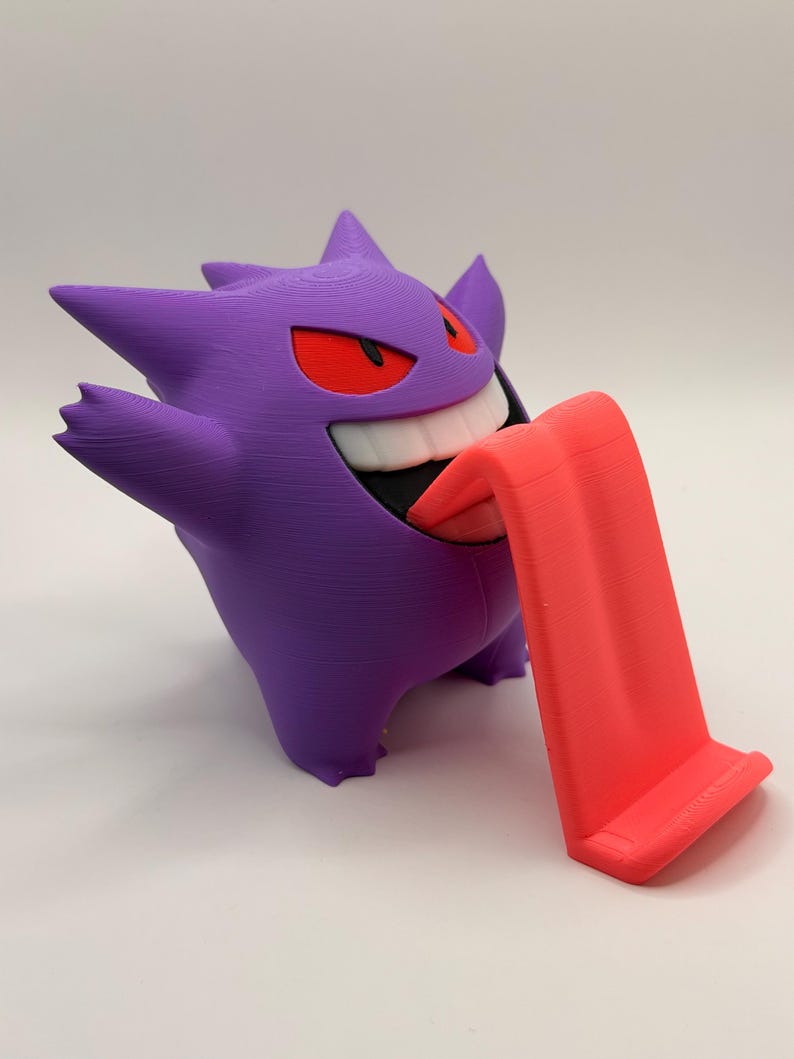 May include: A purple Gengar figure with red eyes and a wide grin, holding out a bright red tongue that serves as a phone or tablet stand. The figure is 3D-printed and stands on a white surface.