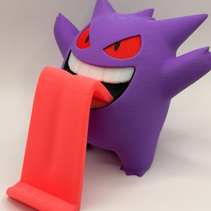 May include: A 3D-printed purple Gengar figure with red eyes and a wide grin, holding out a bright red tongue that serves as a phone or tablet stand. The figure has a textured surface.