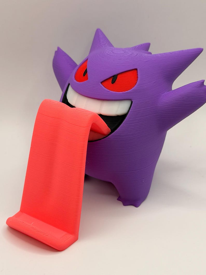 May include: A 3D-printed purple Gengar figure with red eyes and a wide grin, holding out a bright red tongue that serves as a phone or tablet stand. The figure has a textured surface.