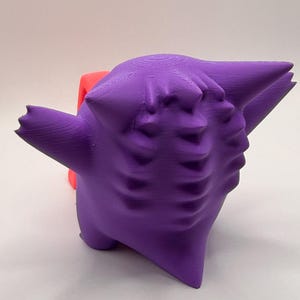 May include: A 3D-printed purple figure with a stylized, cartoonish design. The figure has a rounded body, pointed ears, and a face with multiple indentations. A red object is partially visible behind the purple figure.