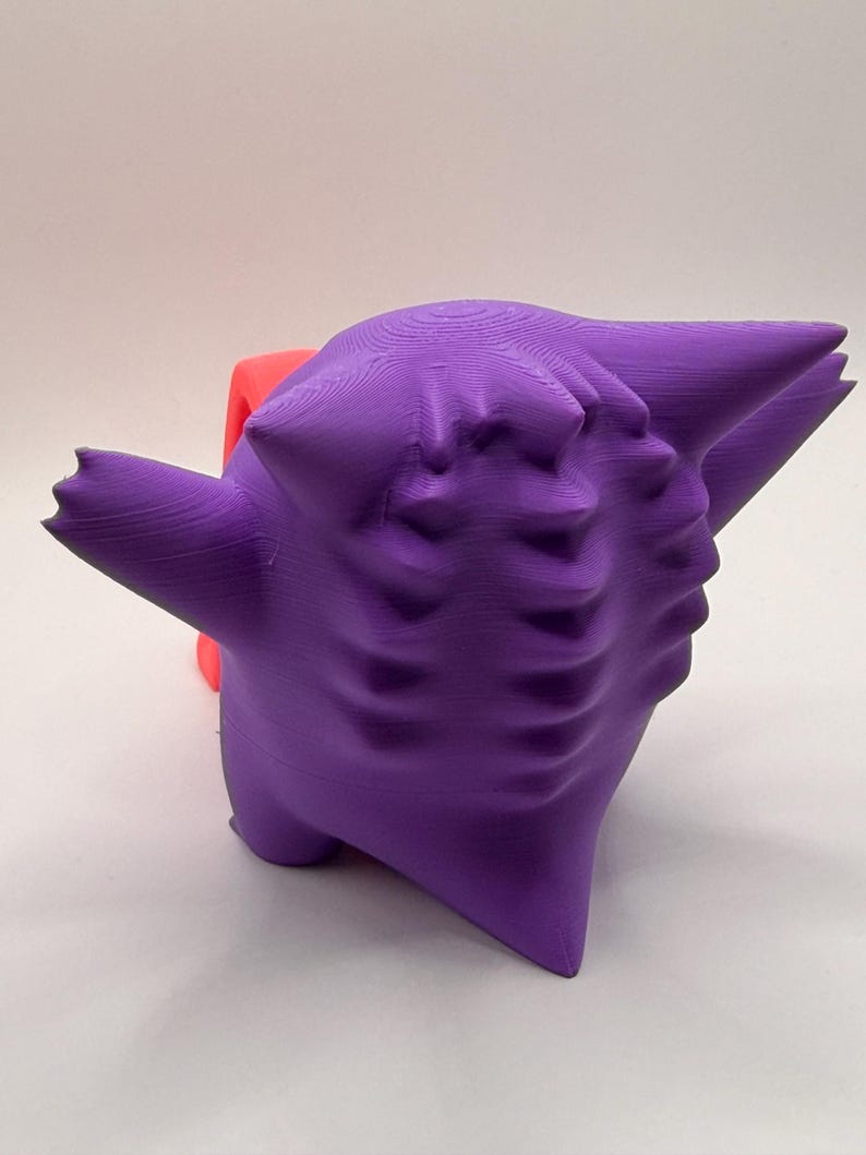 May include: A 3D-printed purple figure with a stylized, cartoonish design. The figure has a rounded body, pointed ears, and a face with multiple indentations. A red object is partially visible behind the purple figure.