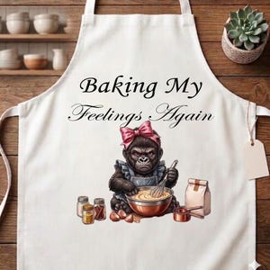 May include: White apron with the text "Baking My Feelings Again." The apron features a cartoon gorilla wearing a pink bow and apron, whisking batter in a bowl. Additional elements include spice jars, eggs, and baking supplies.