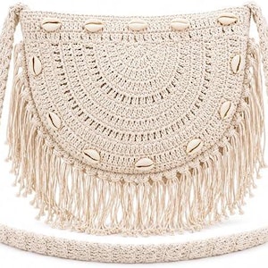 Puede incluir: Un bolso de hombro de ganchillo en color crema con forma semicircular, que presenta un patrón de tejido texturizado, un borde con flecos largos en la parte inferior y una serie de conchas de cauri blancas dispuestas en arco al frente.