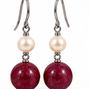 Può includere: Un paio di orecchini pendenti con una grande perla sferica in pietra rosso scuro alla base, sormontata da una perla più piccola, liscia e color crema. Le perle sono collegate da maglie in metallo argentato a un gancio per orecchini.