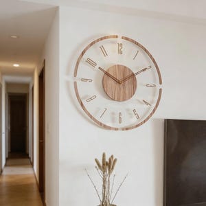 Puede incluir: Un reloj de pared moderno con una esfera transparente y un marco de madera. El reloj presenta un diseño circular con números y manecillas de madera. El centro del reloj es un círculo de madera maciza. El reloj está montado en una pared blanca.