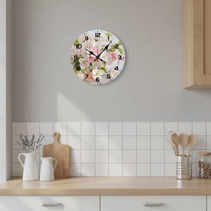 May include: Round wall clock with a floral design featuring white and pink flowers. The clock has black numerals and hands, and is mounted on a light gray wall. The clock is a decorative item for a kitchen or living room.
