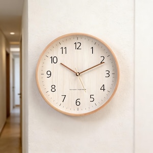 Puede incluir: Un reloj de pared redondo con marco de madera clara y esfera blanca. El reloj tiene números negros, manecillas marrones y un segundero. Está montado en una pared blanca.