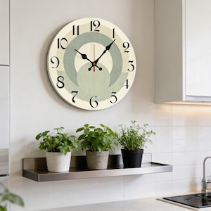 May include: A circular wall clock with a cream face, black numerals, and black hands. The clock has a geometric design in green and grey. Below the clock is a stainless steel shelf with three potted plants.