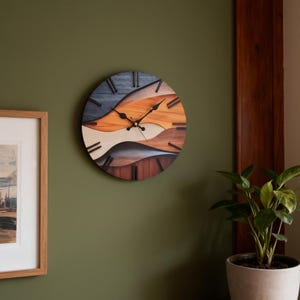 Pode incluir: Um relógio de parede redondo de madeira com um design em camadas. O mostrador do relógio apresenta um padrão ondulado em tons de azul, laranja, creme e castanho. Ponteiros e marcadores de hora pretos completam o design. O relógio está montado numa parede verde.