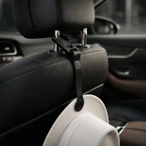 Könnte beinhalten: Ein schwarzer Kopfstützenhaken für das Auto mit einem lederartigen Riemen und einem runden Endstück, das die geprägten Buchstaben AM trägt. Der Haken ist an der Metallstange eines schwarzen Ledersitzes befestigt und hält einen hellen Hut.