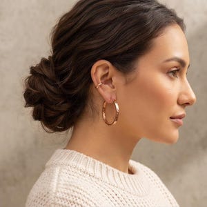 May include: A pair of rose gold hoop earrings with a hammered texture and a matching rose gold ear cuff. The hoop earrings are circular and hang from the earlobe, while the ear cuff is positioned on the upper cartilage of the ear.
