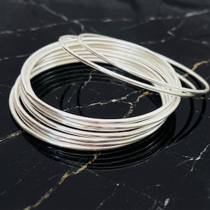 May include: A set of multiple thin, polished silver-colored bangle bracelets stacked together. The bangles are circular, smooth, and reflective, featuring a simple, unadorned design with a consistent metallic finish throughout the entire set.