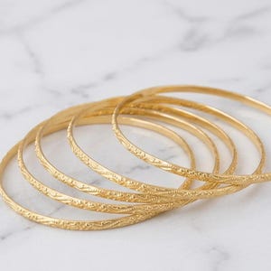 Op de afbeelding: Een set van vijf dunne, ronde gouden armbanden, elk voorzien van een delicaat, herhalend gegraveerd patroon over het gehele buitenoppervlak. De armbanden zijn in een licht overlappende, gestapelde formatie gerangschikt.