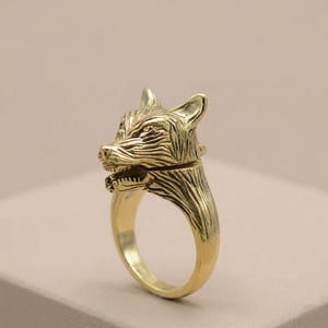 May include: A gold-colored ring featuring a detailed wolf head design. The ring showcases the wolf's face with intricate carved details, including the ears, eyes, and snout. The ring's band is a simple, smooth gold color.