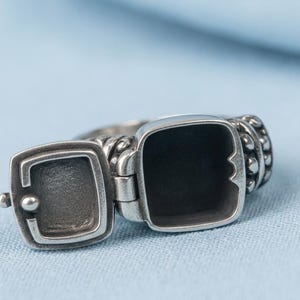 Puede incluir: Un anillo de metal plateado con un compartimento de relicario cuadrado con bisagras que está abierto. La banda del anillo muestra un diseño texturizado con cuentas en los lados, y el interior del relicario es hueco y de apariencia oscura.