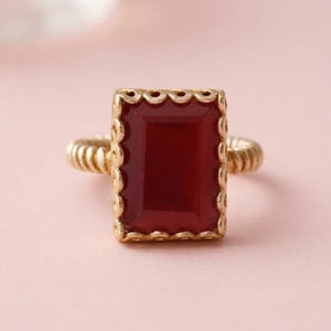 Op de afbeelding: Een gouden ring met een rechthoekige, dieprode steen in een geschulpte, getextureerde gouden zetting. De ringband heeft een opvallend geribbeld of gedraaid ontwerp, wat zorgt voor een textuur die de centrale zetting aanvult.