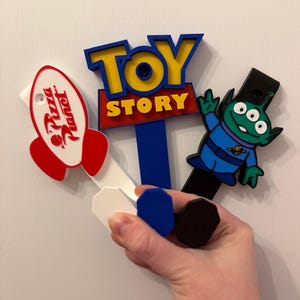 May include: Three decorative wall hooks featuring Toy Story themes. One hook is a white Pizza Planet rocket, one is a blue Toy Story logo sign, and one is a green three-eyed alien character. Each hook has a flat, coloured base for mounting.