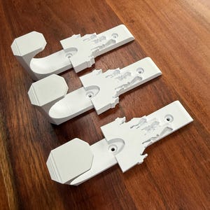 May include: Three white wall hooks with a unique design. Each hook features a rectangular base with a detailed, stylized map-like shape and a curved hook. The hooks are arranged on a dark wood surface.