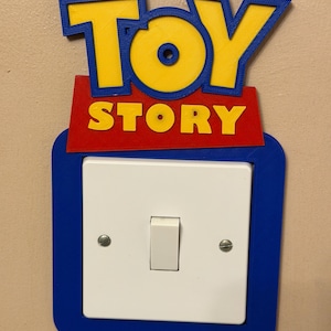 Puede incluir: Una cubierta decorativa para interruptor de luz azul, roja y amarilla con las palabras "TOY STORY" en letras amarillas. La cubierta está diseñada para encajar alrededor de un interruptor de luz estándar y está montada en una pared beige.
