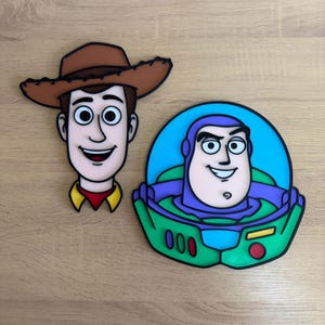 Könnte beinhalten: Zwei flache, dekorative Wandplaketten mit den Zeichentrickfiguren Woody und Buzz Lightyear. Woody trägt einen braunen Cowboyhut, während Buzz in seinem charakteristischen grünen, violetten und blauen Weltraum-Ranger-Anzug dargestellt ist.