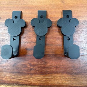 May include: Three black metal wall hooks featuring a Mickey Mouse head design. Each hook has a rectangular top with two screw holes, a Mickey Mouse head, and a curved hook. The hooks are displayed on a wooden surface.
