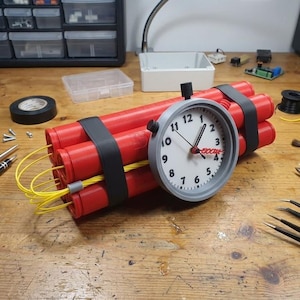 Puede incluir: Un artículo de novedad que se asemeja a la dinamita, con cilindros rojos sujetos con correas negras. Un reloj con un bisel gris está adjunto, mostrando la hora. Cables amarillos se extienden desde los cilindros. El artículo está sobre una superficie de madera con herramientas.