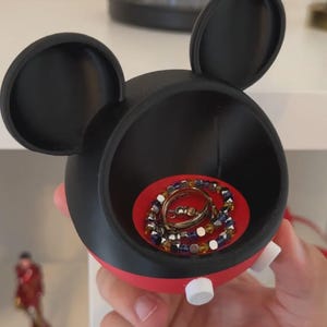 May include: A black and red circular storage container shaped like a mouse head with two large rounded ears. The interior base is bright red and holds a metallic ring and a beaded bracelet with blue, yellow, and silver-toned square beads.