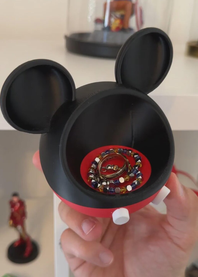 May include: A black and red circular storage container shaped like a mouse head with two large rounded ears. The interior base is bright red and holds a metallic ring and a beaded bracelet with blue, yellow, and silver-toned square beads.