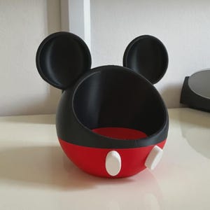 May include: A spherical black and red container featuring two large circular black ears on top. The lower portion is red with two white oval accents on the front. The interior is black, and the top section is open, creating a bowl-like shape.