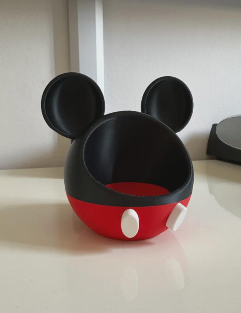 May include: A spherical black and red container featuring two large circular black ears on top. The lower portion is red with two white oval accents on the front. The interior is black, and the top section is open, creating a bowl-like shape.