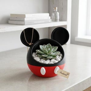 May include: A black and red spherical planter with two circular ears, featuring two white oval accents on the front. The planter contains a green succulent nestled in white pebbles and holds a gold chain. A small beige tag reads Handmade.
