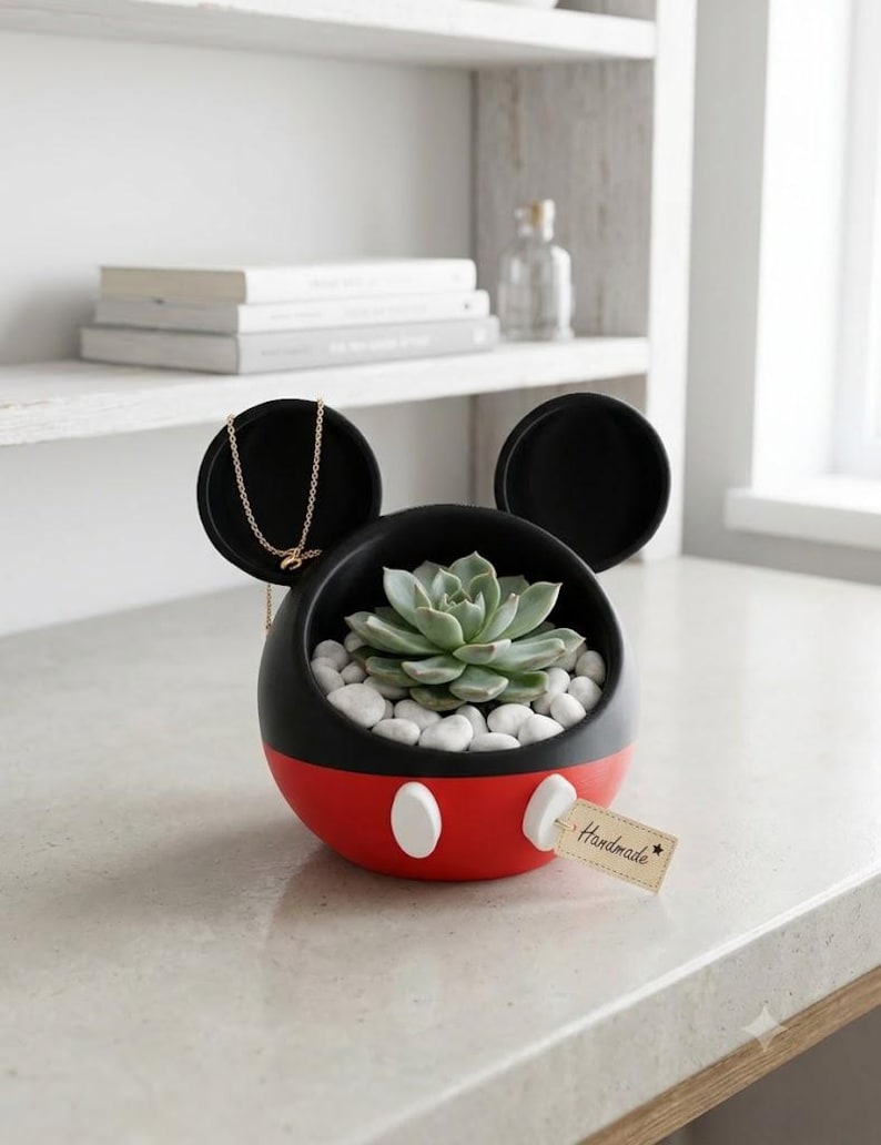 May include: A black and red spherical planter with two circular ears, featuring two white oval accents on the front. The planter contains a green succulent nestled in white pebbles and holds a gold chain. A small beige tag reads Handmade.